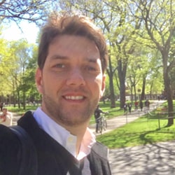 Homem branco com topete e sorrindo, com camisa social branca e suéter preto em um parque com árvores.