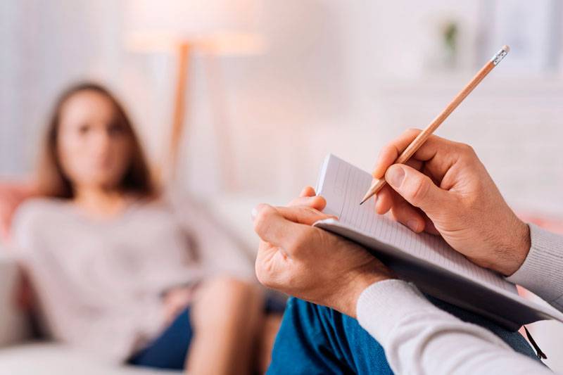 Homem segurando um lapis e um caderno simulando escrever e de fundo uma mulher desfocada e sentada como se estivesse em uma consulta.