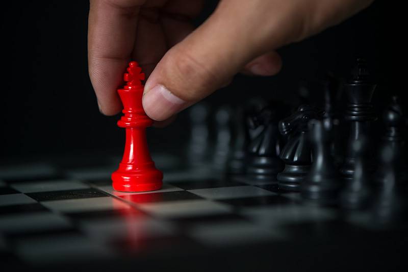 Imagem de uma mão segurando uma peça vermelha de xadrez e outras peças de xadrez na cor preta de fundo.