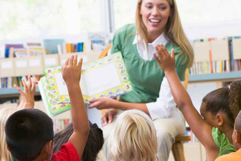 Imagem de uma mulher loira apresentando um livro para crianças e as crianças com as mãos levantadas.